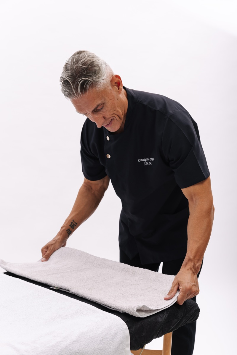 Juan Manuel Morales in a black osteopath’s tunic, spreading a towel over the treatment table