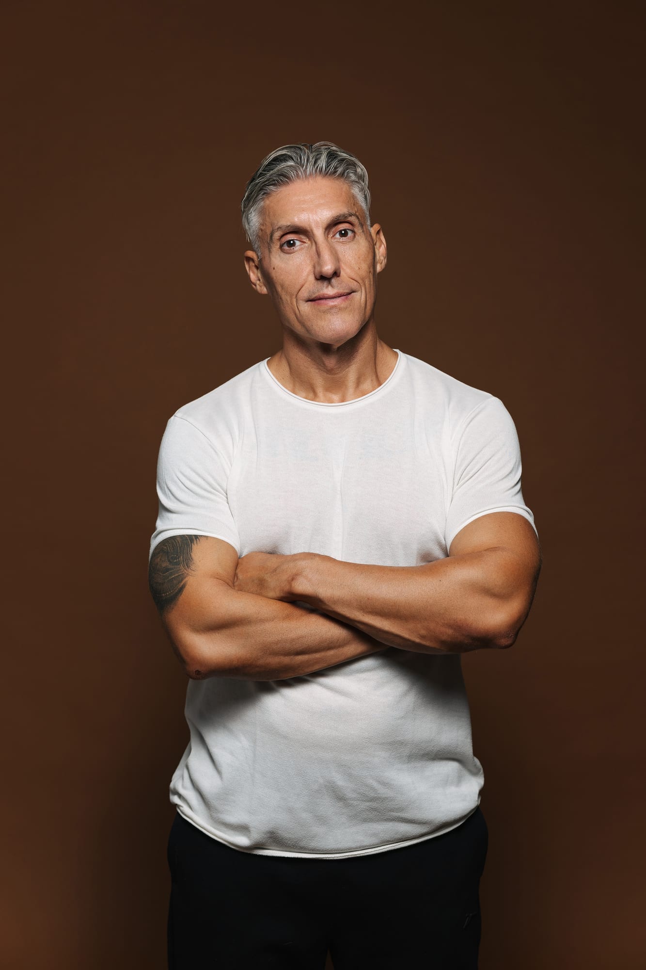 Juan Manuel Morales in a white T-shirt with his arms crossed, looking at the camera against a warm brown background