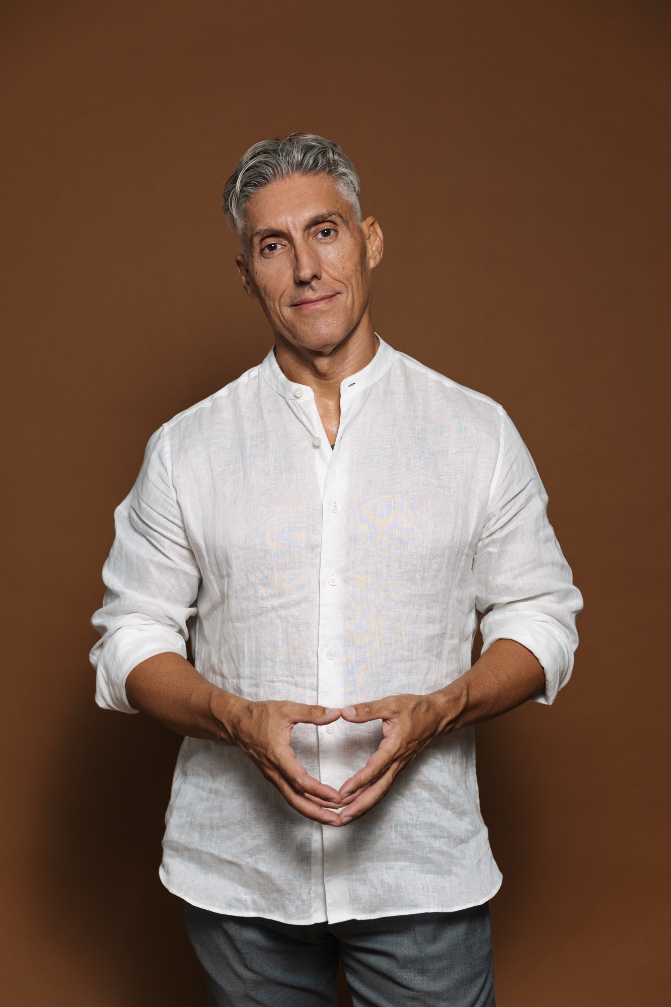 Juan Manuel Morales con camisa de lino blanca sobre fondo marrón cálido, de pie y con las manos juntas a la altura de la cintura