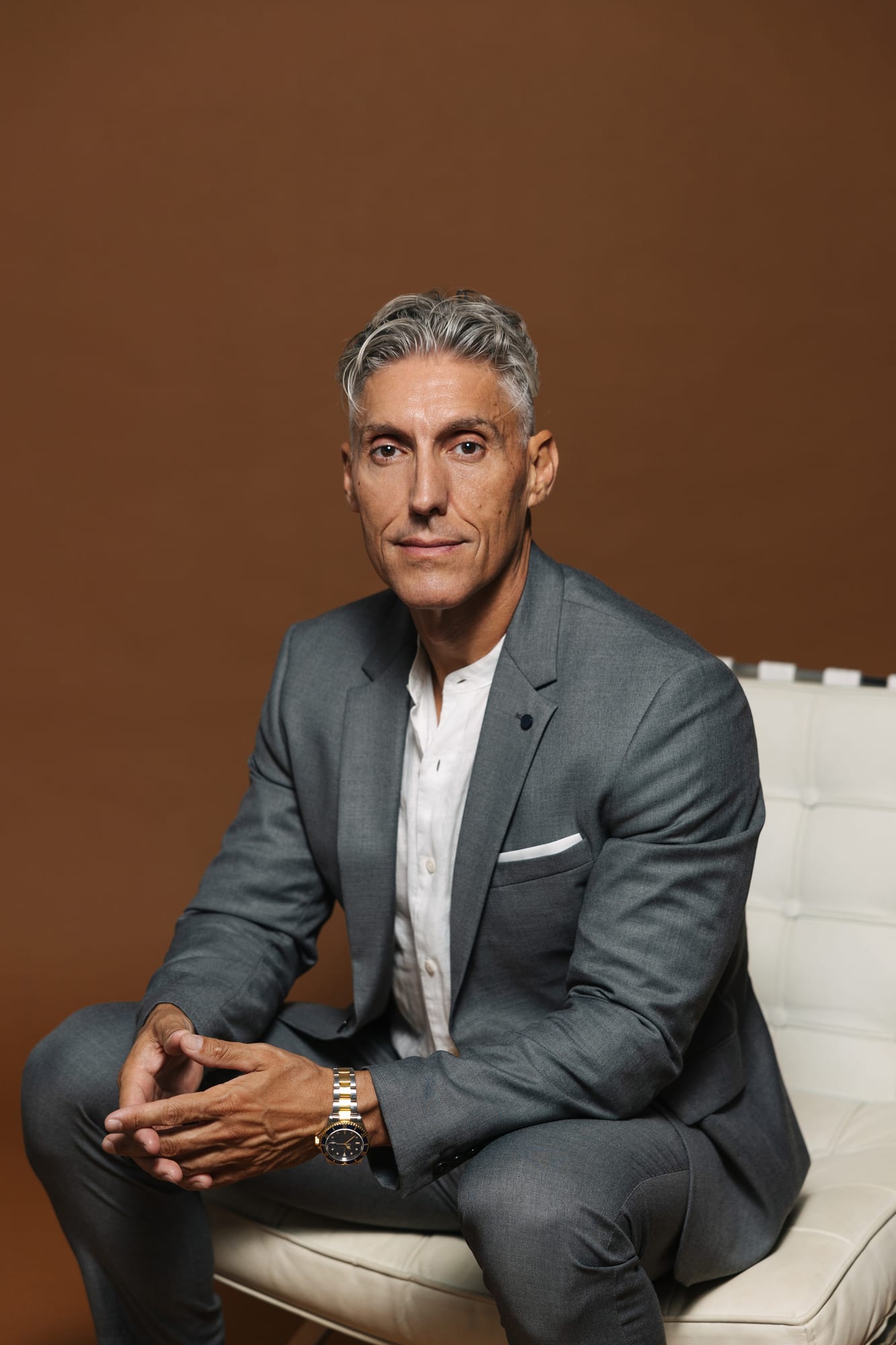 Juan Manuel Morales in a grey suit, seated on a white bench with his hands clasped, looking at the camera against a warm brown background
