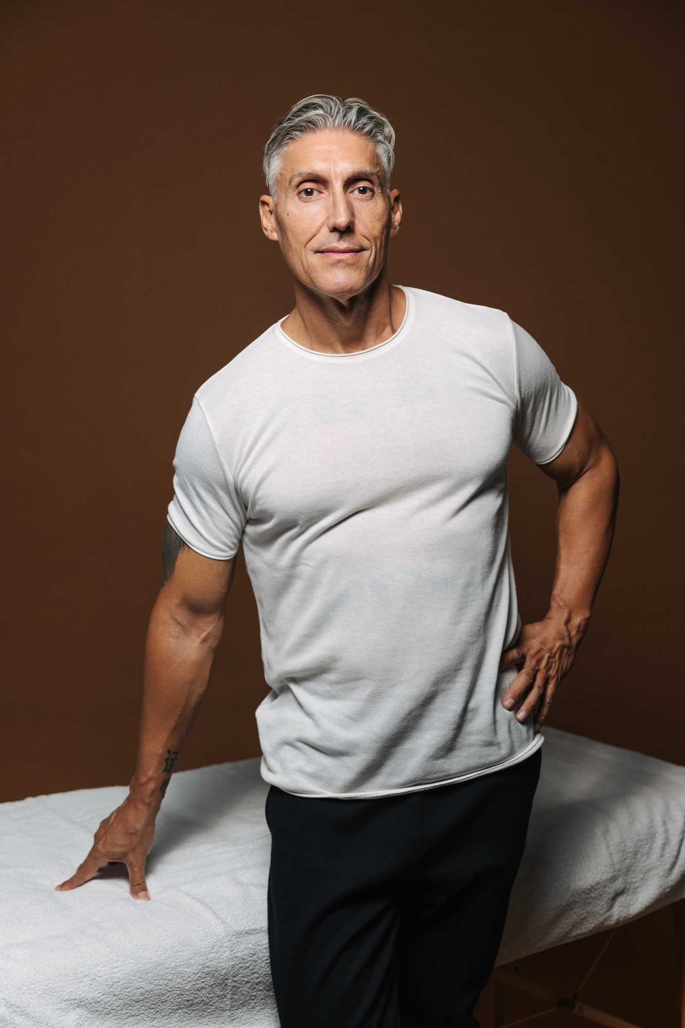 Juan Manuel in a white T-shirt and black training trousers, one hand resting on the treatment table, against a brown backdrop