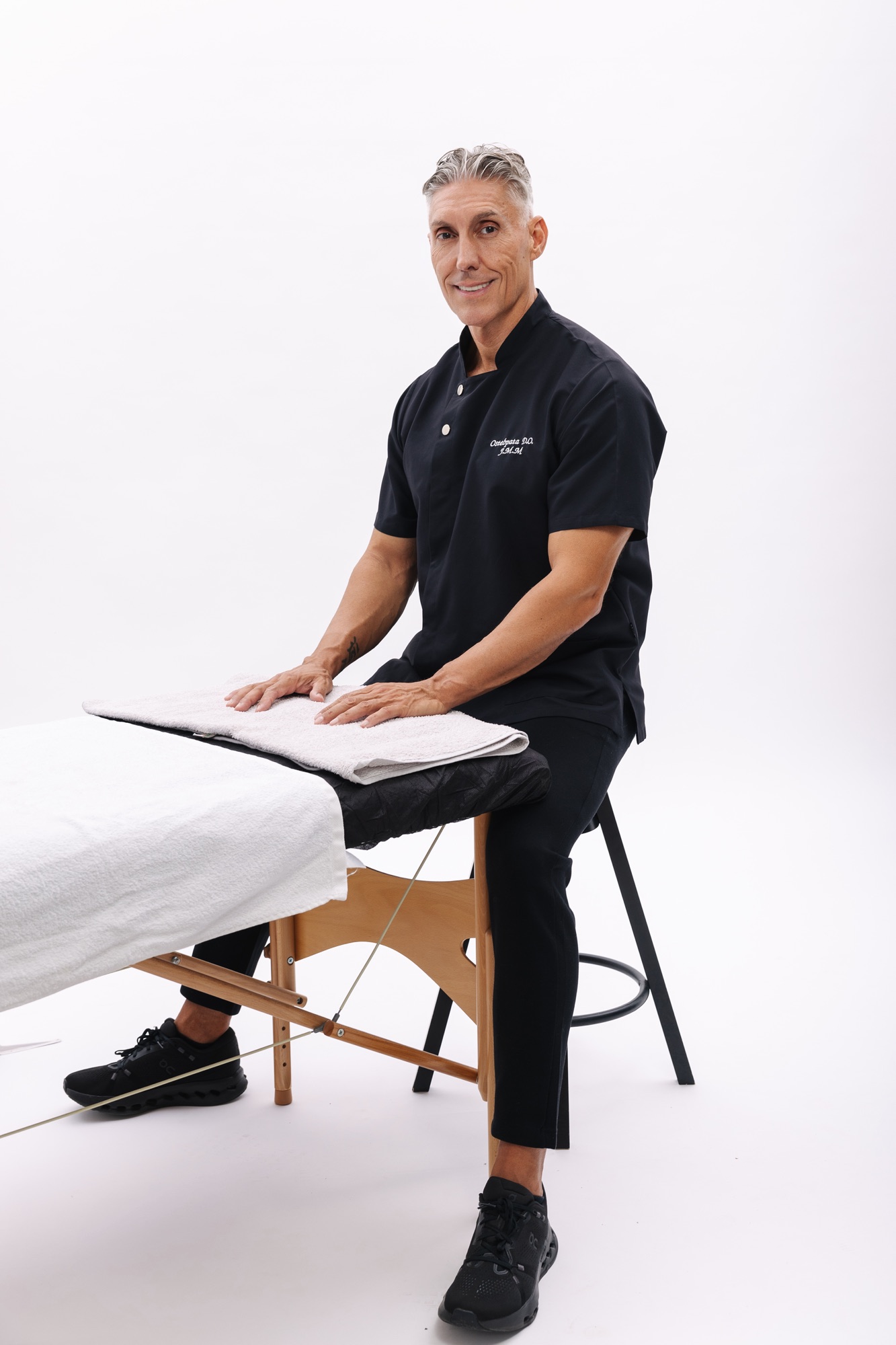 Juan Manuel Morales in a black osteopath’s tunic, seated on a stool beside the treatment table, hands resting on the towel