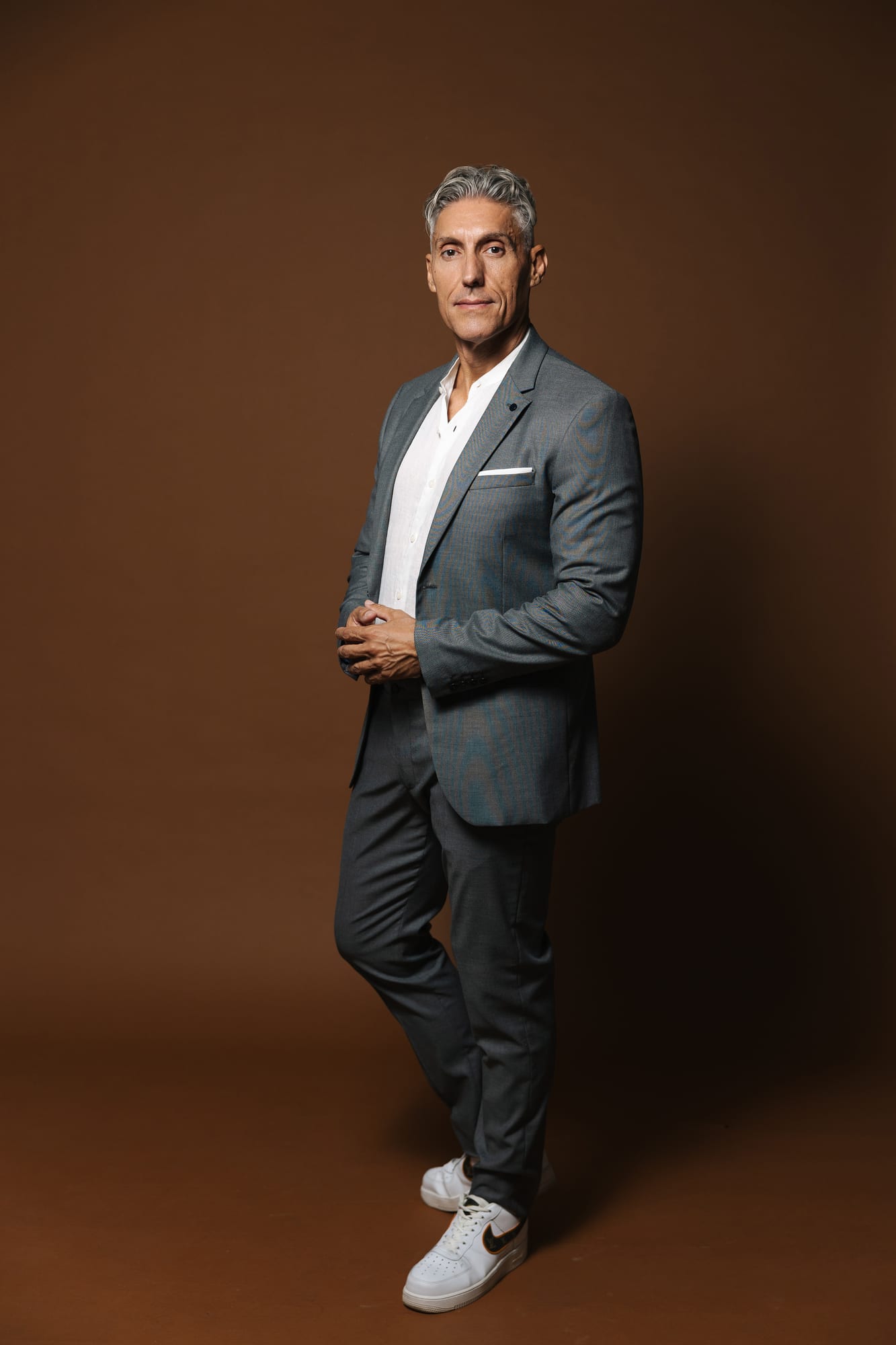 Juan Manuel Morales in a grey suit and white trainers, full length and mid-stride, against a warm brown background