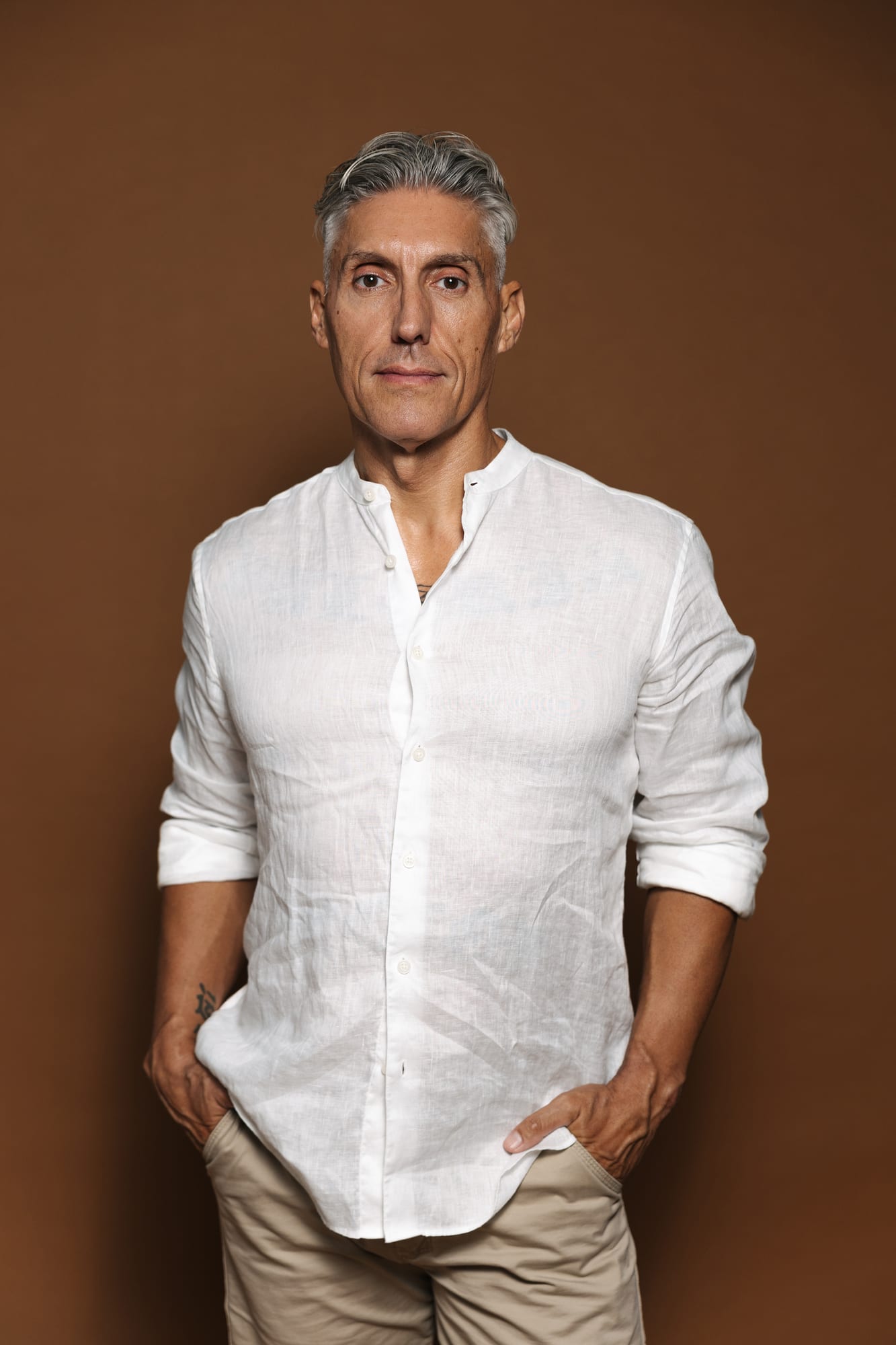 Juan Manuel Morales con camisa de lino blanca y pantalón beige, de pie con las manos en los bolsillos, sobre fondo marrón cálido