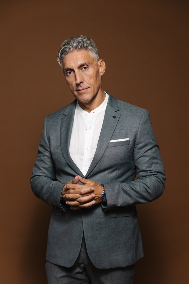 Retrato de Juan Manuel en traje gris, manos entrelazadas, sobre fondo marrón cálido