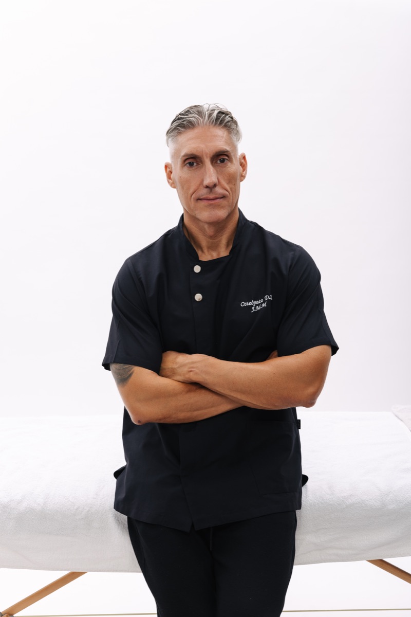 Juan Manuel Morales in a black osteopath’s tunic, arms crossed, standing in front of a treatment table